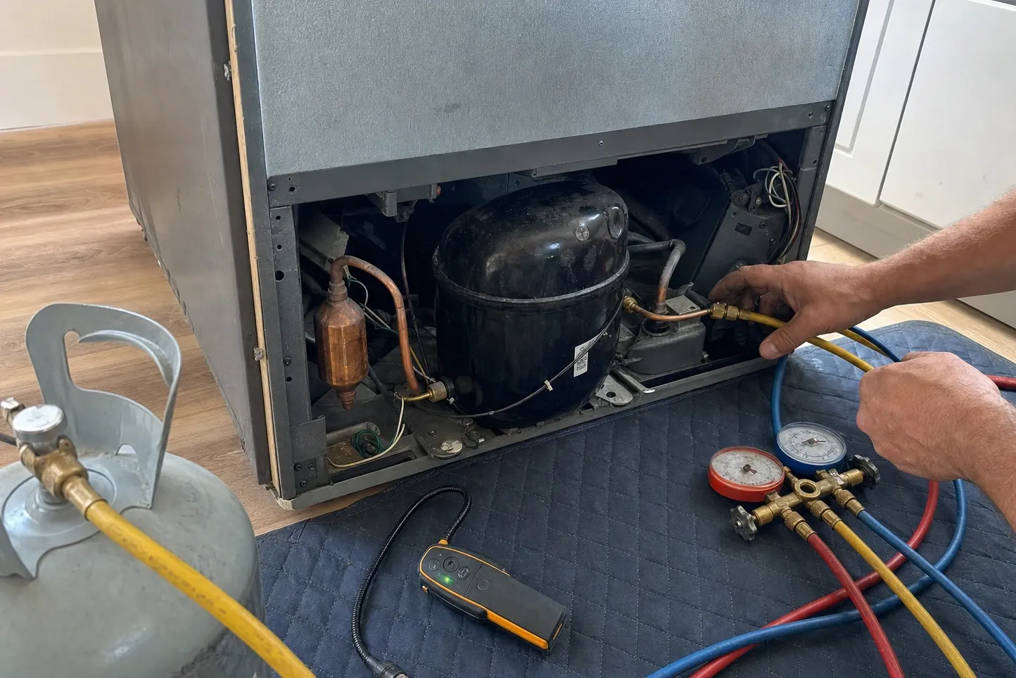 Refrigerator pulled out from the wall with its rear panel off and a charging hose connected at the compressor, manifold gauges and a refrigerant cylinder on the floor